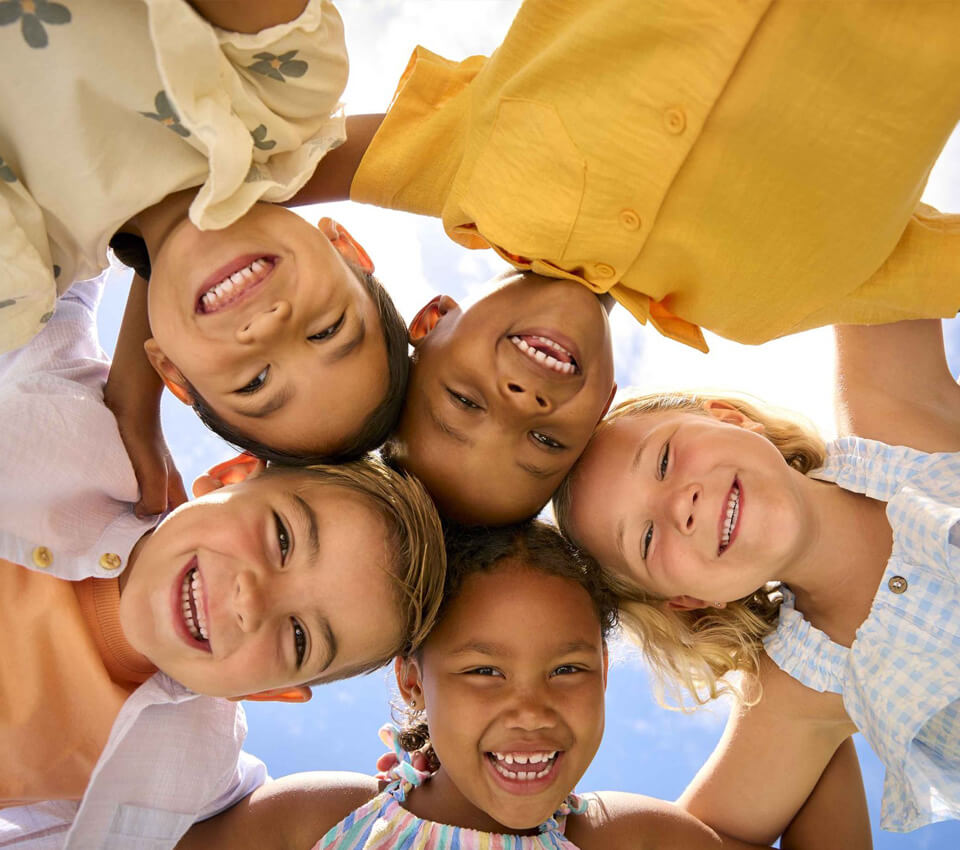 Five preschool children huddled together, smiling down at the ground on a sunny day.
