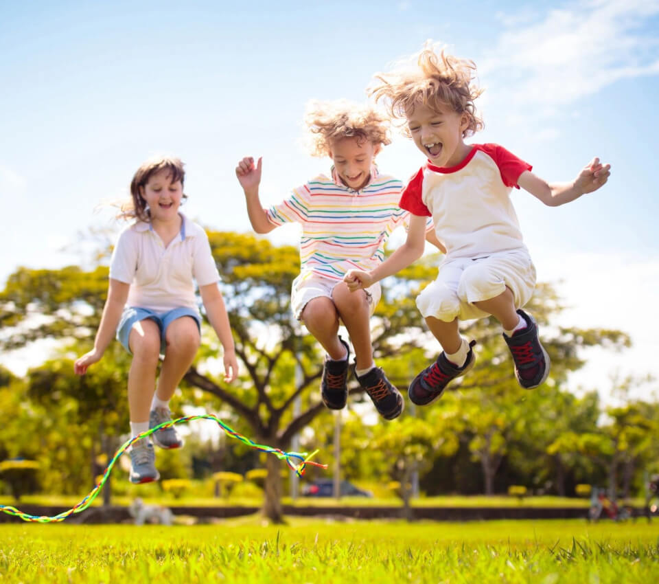 Three children jumping on a green grassy field under bright sunlight on a clear day.