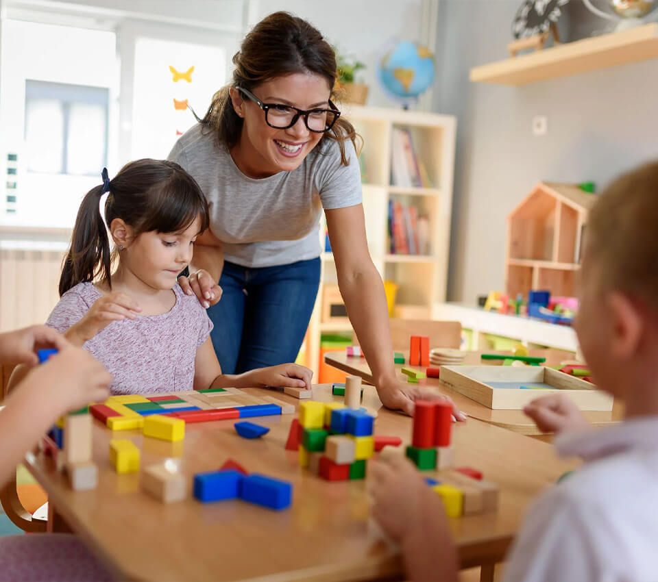 Preschool teacher smiling while observing children playing with colorful educational blocks at a classroom table.